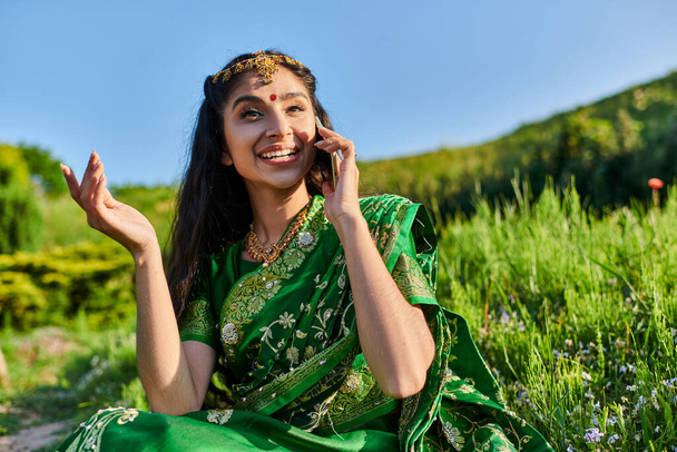 smiling young indian woman in green sari talking on smartphone while sitting on grass in summer - 写真・画像