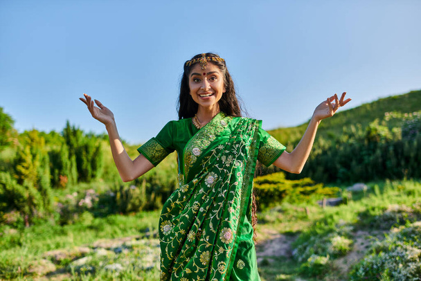 cheerful young indian woman in green sari posing and looking at camera on summer field - Fotó, kép
