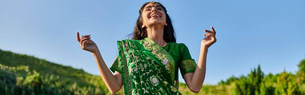 щаслива молода індійська жінка в зеленому сарі, що стоїть з літнім пейзажем на фоні, банер - Фото, зображення