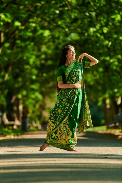 volle Länge der stilvollen und lächelnden indischen Frau in Sari posiert auf der Straße mit Bäumen im Hintergrund - Foto, Bild