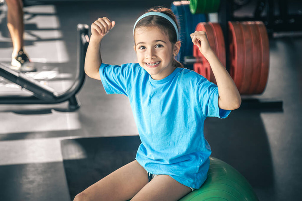 Charming little girl is shows her biceps and smiling while working out at gym. - Photo, Image
