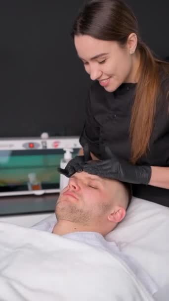 Un uomo sdraiato su un letto in una spa riceve un massaggio facciale della pelle con olio essenziale aromatico cura della pelle. Video verticale - Filmati, video
