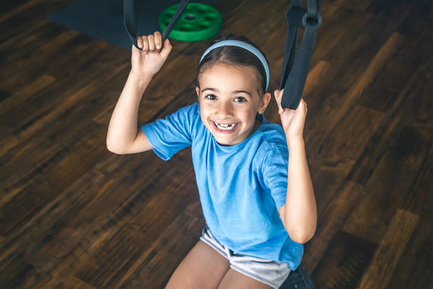 Bambina che si allena al chiuso utilizzando cinghie da fitness trx per lavorare con il proprio peso, facendo squat o allenamento parte superiore del corpo. - Foto, immagini