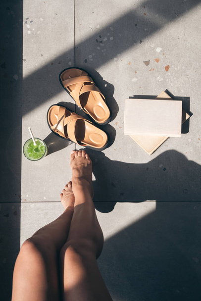Літня композиція з тапочками, лимонадом, книгою та жіночими ногами на плитці з тінями на сонячний день, вид зверху. - Фото, зображення