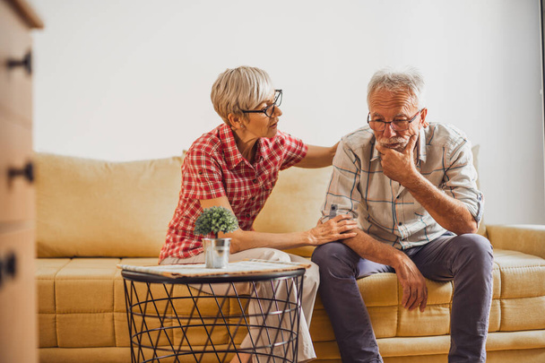 Senior couple sitting in living room. Man is sad and worried and woman is consoling him. - Photo, Image