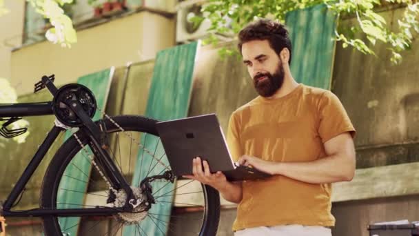 Здоровый преданный кавказский мужчина хватается за свой ноутбук, чтобы починить сломанный современный велосипед с помощью онлайн-руководства. Любящий спорт мужчина-велосипедист держит мини-компьютер с инструкциями по уходу за велосипедом на открытом воздухе. - Кадры, видео