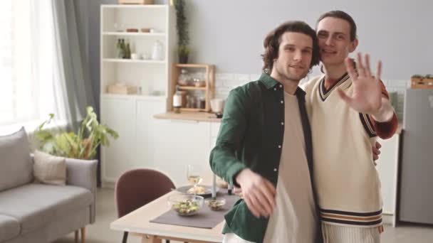 Retrato lento de feliz pareja gay caucásica sonriendo y saludando a la cámara de pie en la acogedora cocina con mesa de cena servida para dos - Imágenes, Vídeo