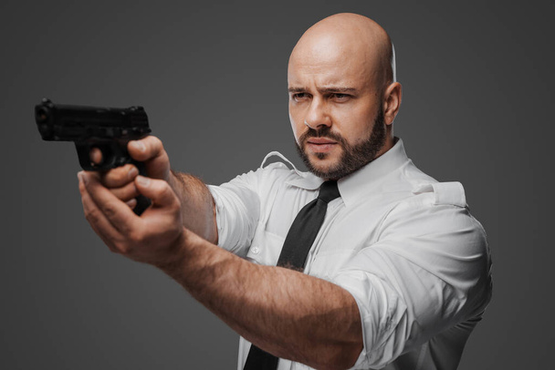 Kaal, man met baard in wit shirt en stropdas richt een pistool, kanaliseert een detective of bodyguard vibe tegen een grijze studio achtergrond - Foto, afbeelding