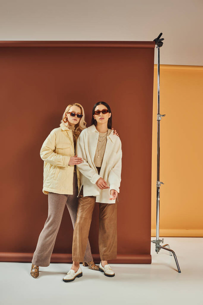 fall season, interracial women in sunglasses and outerwear posing together on duo color backdrop - Photo, Image