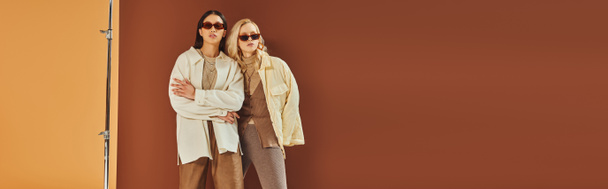 fall trends, interracial women in sunglasses and outerwear posing on duo color backdrop, banner - Photo, Image