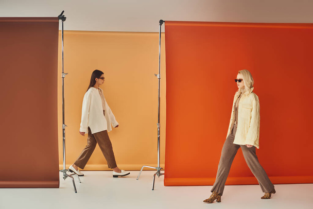 πολυπολιτισμικές γυναίκες το φθινόπωρο εξωτερικά ενδύματα και γυαλιά ηλίου περπάτημα κοντά πολύχρωμο φόντο, φθινοπωρινή μόδα - Φωτογραφία, εικόνα