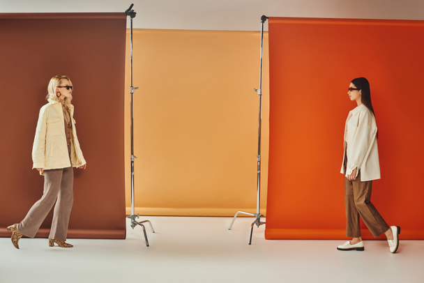 διαφυλετικά μοντέλα σε εξωτερικά ενδύματα και γυαλιά ηλίου περπάτημα κοντά σε πολύχρωμο φόντο, πτώση έννοια της μόδας - Φωτογραφία, εικόνα
