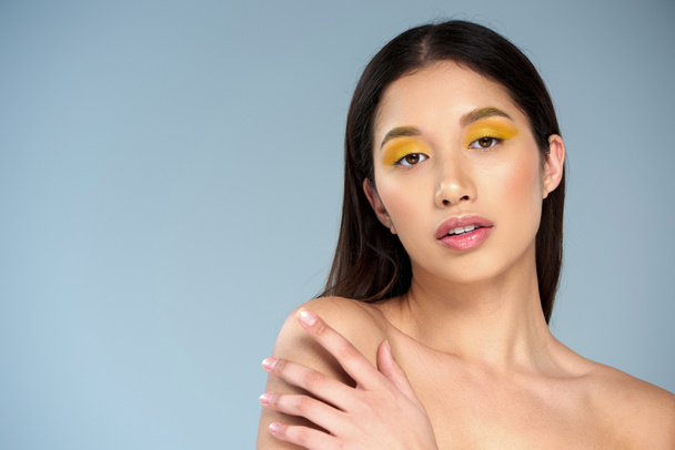 zelfexpressie, jong aziatisch model met gedurfde make-up en blote schouders poserend op een blauwe achtergrond - Foto, afbeelding