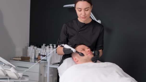 Gezichtsreiniging. Procedure voor het ultrasone apparaat voor het gezicht. Behandeling van acne bij mannen. Een man krijgt een elektrische hijsmassage in een spa. Verticale video - Video