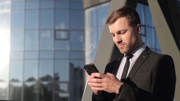 Un joven hombre de negocios sonriente con un traje negro está mirando un teléfono inteligente. Gerente cerca del centro de negocios con un teléfono móvil en sus manos. Concepto empresarial - Imágenes, Vídeo