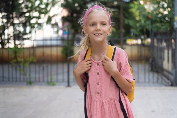 Blondi koulutyttö hymyilee tilalla hihnat reppu söpö pieni tyttö odottaa vanhempia oppituntien jälkeen seisoo kadulla koulun lähellä lämpimänä syyspäivänä - Valokuva, kuva
