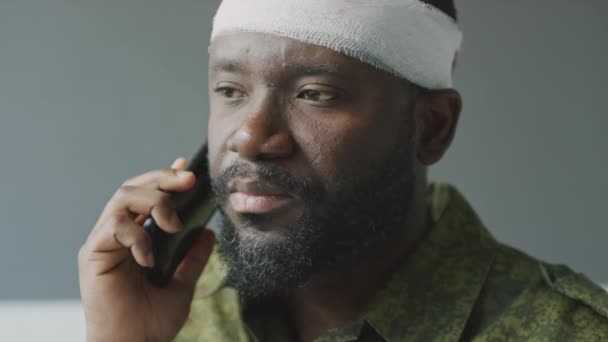 Zbliżenie portret brodatego Afroamerykanina kapitana z urazem głowy siedzącego w pokoju szpitalnym rozmawiającego przez telefon - Materiał filmowy, wideo
