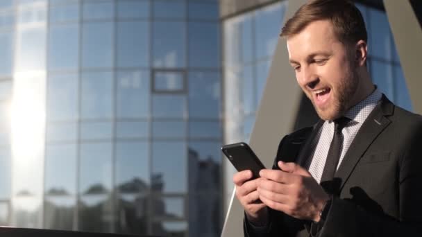Nuori liikemies lähellä liikekeskusta matkapuhelin kädessään. Nuori hymyilevä liikemies mustassa puvussa katselee älypuhelinta. Maailman yrityskonsepti - Materiaali, video