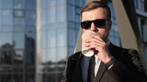 Νεαρός επιχειρηματίας με γενειάδα και μαύρα γυαλιά πίνει καφέ έξω σε μια όμορφη ηλιόλουστη μέρα. Διευθυντής στον ουρανοξύστη γυαλιού υποβάθρου - Πλάνα, βίντεο