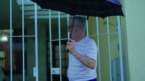 Ανώτερος άντρας ανοίγει ομπρέλα για προστασία από τη βροχή, βγαίνει στο δρόμο από το σπίτι. Ηλικιωμένοι εγκαταλείπουν την κατοικία τους, ανοίγει την πύλη περπατά στο αστικό πεζοδρόμιο ενώ βρέχει - Φωτογραφία, εικόνα