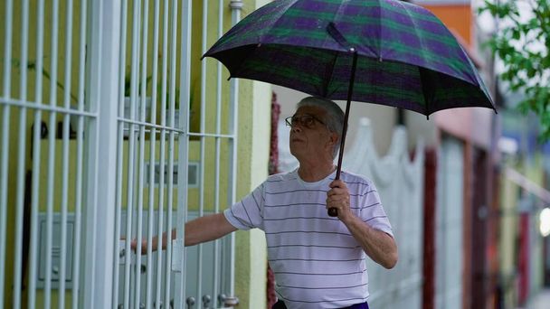 Hombre mayor que regresa a casa bajo la lluvia, despliega paraguas y asegura la puerta, camina por la acera - Foto, imagen