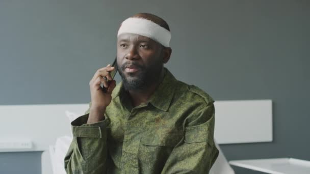 Militar afroamericano con herida de bala usando camuflaje sentado en la habitación del hospital hablando por teléfono con su familia o amigos - Imágenes, Vídeo