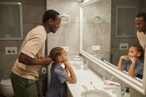 Side view muotokuva huolehtiva musta isä punos tyttäret hiukset peili kylpyhuoneessa ja auttaa saada valmis aamulla - Valokuva, kuva
