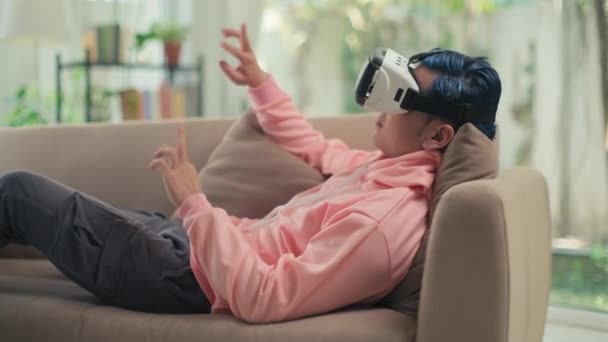 Zoomer με μπλε μαλλιά σε ροζ κουκούλα ξαπλωμένος στον καναπέ με γυαλιά VR και παίζοντας βιντεοπαιχνίδι ενώ ξεκουράζεται στο σπίτι - Πλάνα, βίντεο