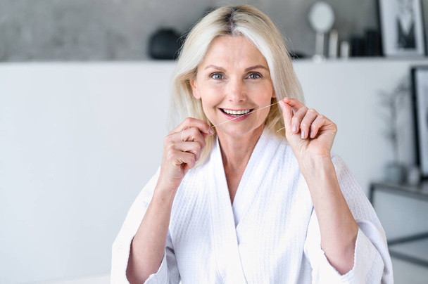 Портрет 40-летней улыбающейся здоровой женщины с зубной нитью в руках. Гигиена и пероральный уход. Уборка, отбеливание. Личное здравоохранение. Утренняя рутина в ванной - Фото, изображение