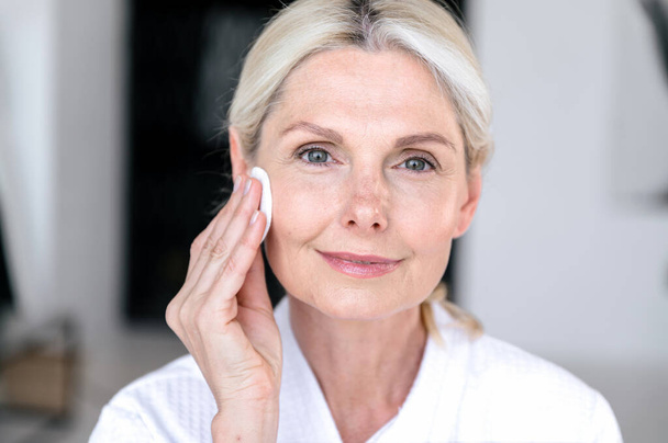 Usuwanie makijażu wodą micelarną i wacikami. 40-letnia kobieta robi codzienną pielęgnację skóry, czyszcząc skórę twarzy naturalnym tonikiem lub balsamem. Portret z bliska. - Zdjęcie, obraz