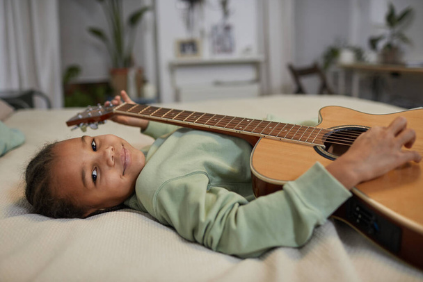 Πορτρέτο του χαριτωμένο μαύρο κορίτσι κρατώντας κιθάρα και κοιτάζοντας κάμερα, ενώ βρίσκεται στο κρεβάτι στο σπίτι, αντίγραφο χώρο - Φωτογραφία, εικόνα