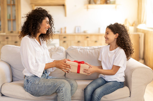Surprise for mom. Happy latin girl giving present box to mommy at home, congratulating with birthday or mothers day, sitting on sofa in living room - Photo, Image