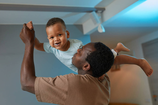 Portret szczęśliwego czarnego ojca bawiącego się słodkim dzieckiem w domu i trzymającego uśmiechniętego syna w powietrzu - Zdjęcie, obraz