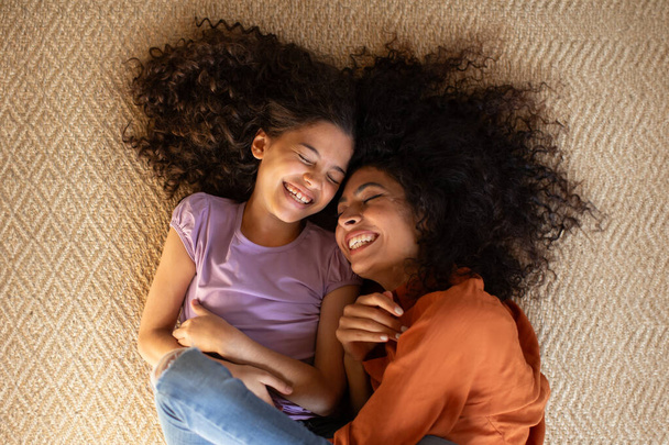 Vista dall'alto della felice madre latina e della sua graziosa figlia sdraiata sul tappeto del pavimento, mamma che abbraccia la ragazza e ride insieme - Foto, immagini