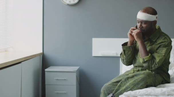 militaire noir avec blessure à la tête assis sur le lit dans la chambre d'hôpital militaire parler au téléphone avec sa famille ou ses amis - Séquence, vidéo