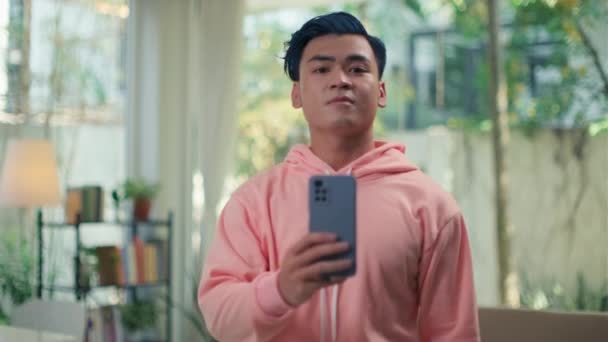 Plan moyen de jeune blogueur asiatique aux cheveux bleus en sweat à capuche rose prenant selfie sur smartphone en miroir pour son réseau social - Séquence, vidéo