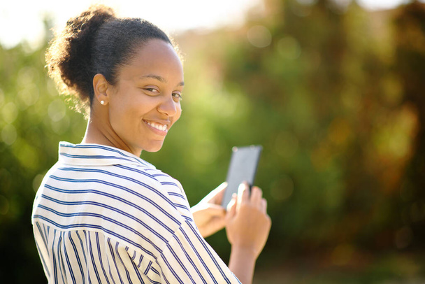 Happy black woman looks at you holding phone in a park - Фото, изображение