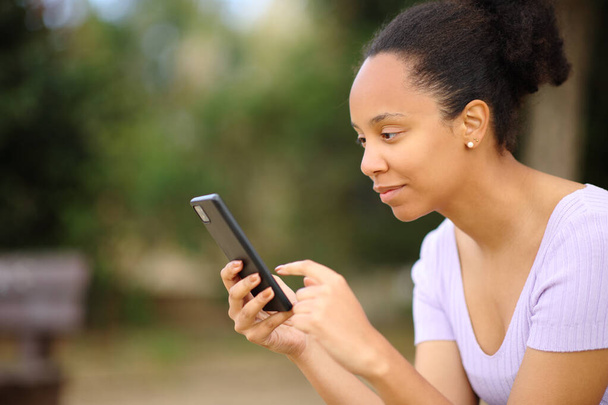 Profile of a black woman using smart phone sitting in a park - Фото, изображение