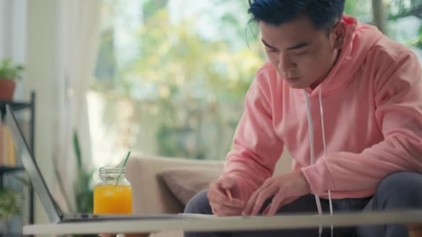 Mladý IT specialista s modrými vlasy pracuje na notebooku z domova, zatímco sedí u stolu s čerstvým nápojem - Záběry, video