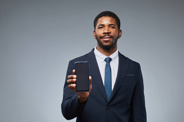 Ritratto di uomo d'affari nero sorridente che tiene in mano il modello di smartphone vuoto e mostra alla fotocamera con sfondo grigio, copia lo spazio - Foto, immagini