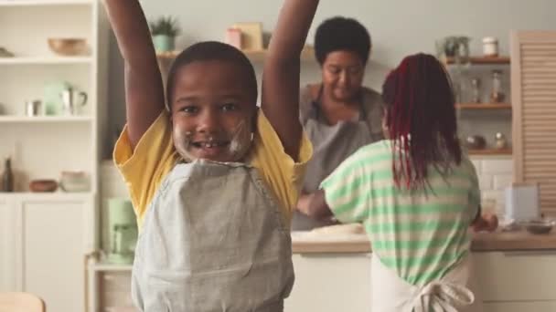 Ritratto del piccolo ragazzo afroamericano con farina sul viso e grembiule in posa per la macchina fotografica mentre aiuta nonna e sorella maggiore in cucina - Filmati, video