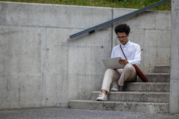 Working remotely. Curly-haired young man in white shirt sitting on the steps with laptop - Fotoğraf, Görsel