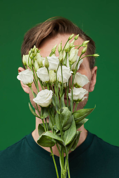 Κάθετο πορτρέτο του ανθρώπου που καλύπτει το πρόσωπο με λευκά τριαντάφυλλα σε ζωντανό πράσινο φόντο - Φωτογραφία, εικόνα