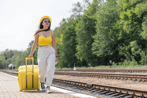 Er wacht een wereld van levendige indrukken: een jonge vrouw met een zonnebril en stijlvolle kleding staat klaar om op reis te gaan. Een leuke en energieke reis: een meisje in een heldere stijlvolle outfit is klaar om te ontdekken - Foto, afbeelding