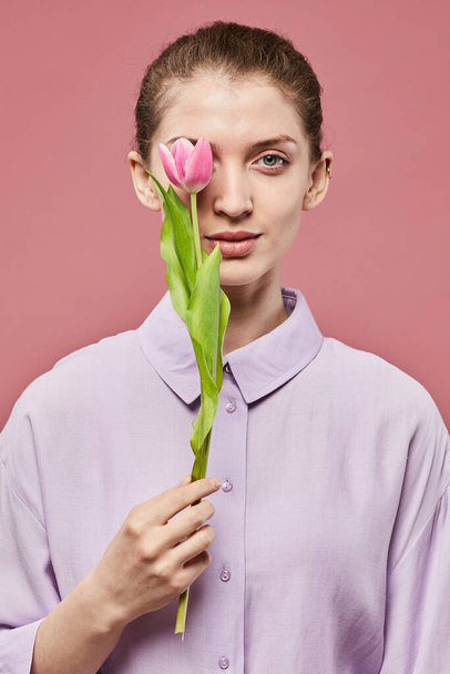 Portrait minimal de jeune femme souriante tenant une seule tulipe rose tout en se tenant debout sur un fond rose vibrant - Photo, image
