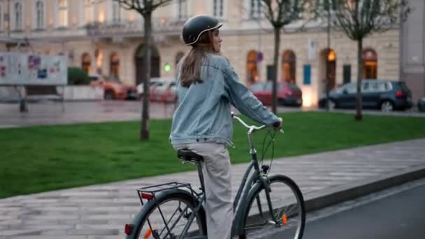 Επιστροφή άποψη του κοριτσιού με κράνος ασφαλείας ποδήλατο ιππασίας σε βροχερή πόλη βράδυ. Νεαρή γυναίκα απολαμβάνει την φιλική προς το περιβάλλον μετακίνηση της στο γραφείο - Πλάνα, βίντεο