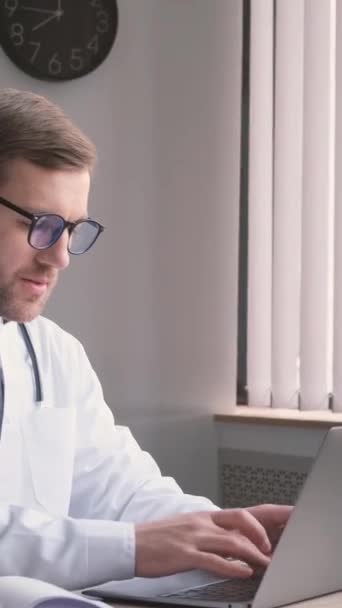Ένας νεαρός ελκυστικός νευρολόγος γιατρός κάθεται στο γραφείο και εργάζεται στον υπολογιστή. Ένας νεαρός ειδικευόμενος δουλεύει σε ένα βιβλιάριο δικτύου. Κάθετη βίντεο - Πλάνα, βίντεο