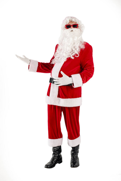 kerstman in rood pak met baard in bril toont zijn handen naar de zijkant en adverteert kopieerruimte op witte geïsoleerde achtergrond, het concept van het nieuwe jaar en kerst - Foto, afbeelding