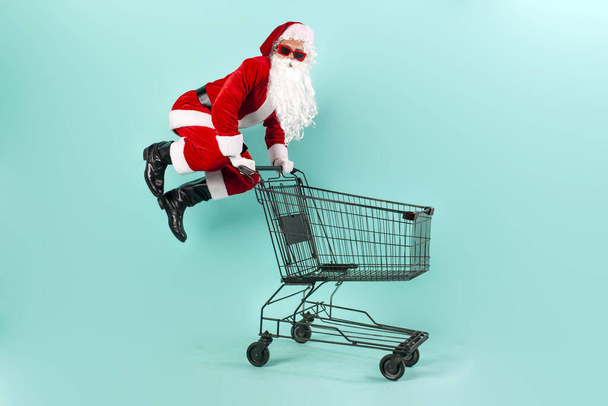 Joulupukin shopper aurinkolaseissa ja puku hyppää ja lentää tyhjällä ostoskorilla myymälään sinisellä eristetyllä taustalla, joulupukki ryntää supermarkettiin ostamaan uutta vuotta ja joulua - Valokuva, kuva