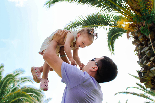 Ένας νεαρός πατέρας κρατά την κορούλα του από πάνω του στο φόντο των φοινίκων. Έννοια της ευτυχισμένης παιδικής ηλικίας, την πατρότητα και ευτυχισμένη οικογένεια. Έννοια του τουρισμού και της αναψυχής. - Φωτογραφία, εικόνα